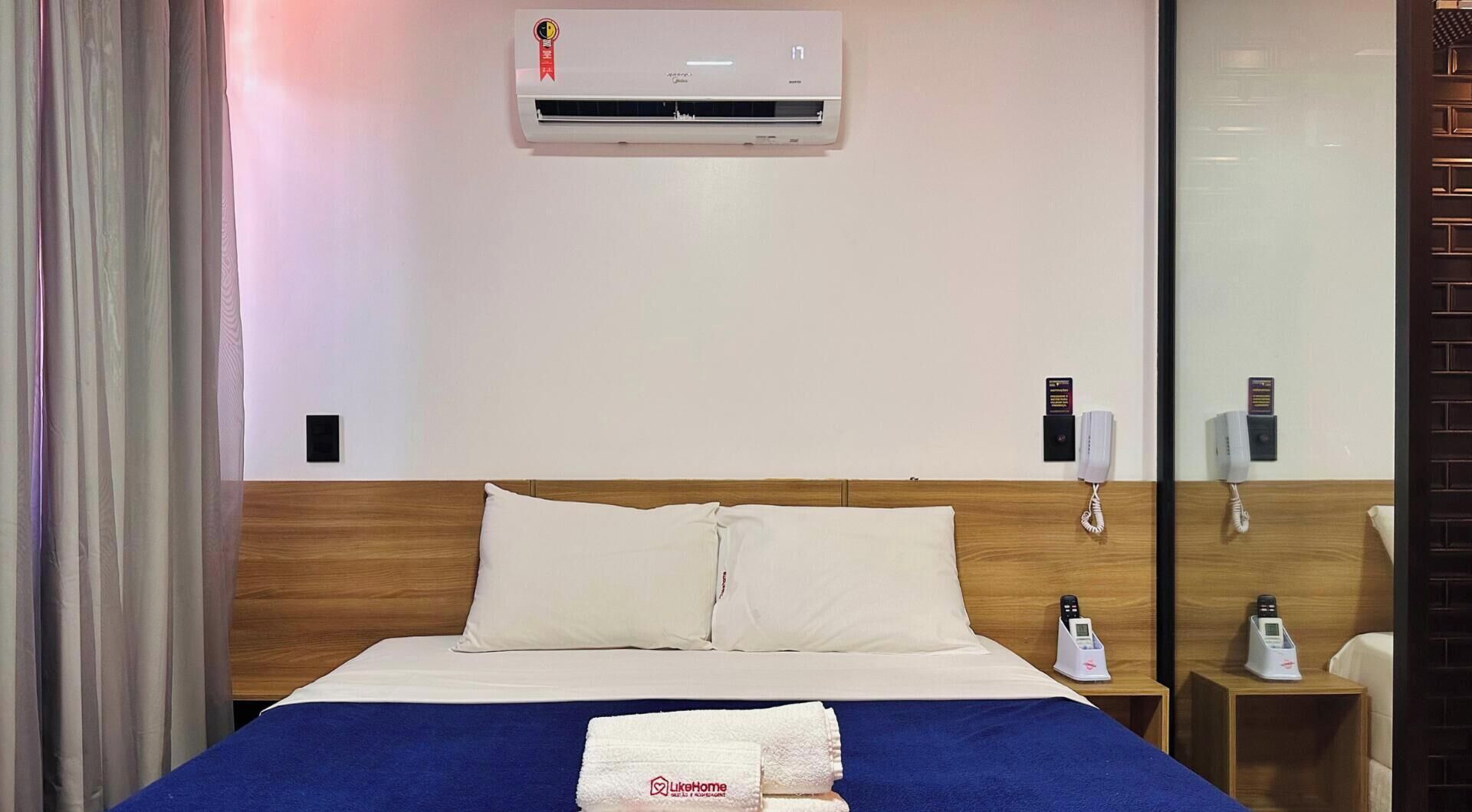 A hotel room with a bed, a bedside table with a phone, a wall-mounted air conditioner, and a glass partition.