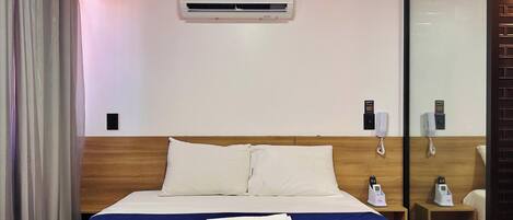 A hotel room with a bed, a bedside table with a phone, a wall-mounted air conditioner, and a glass partition.