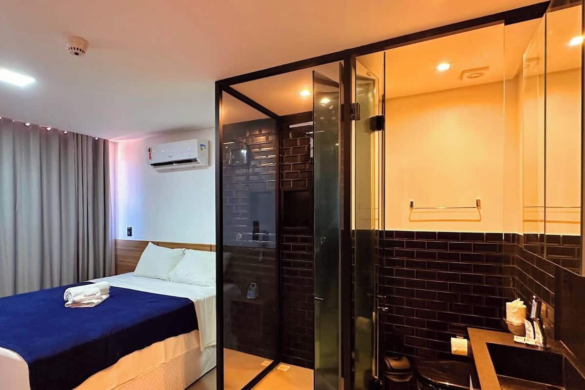 A modern hotel room with a bed, a glass partition, a bathroom with a towel rack, and a small table with toiletries.