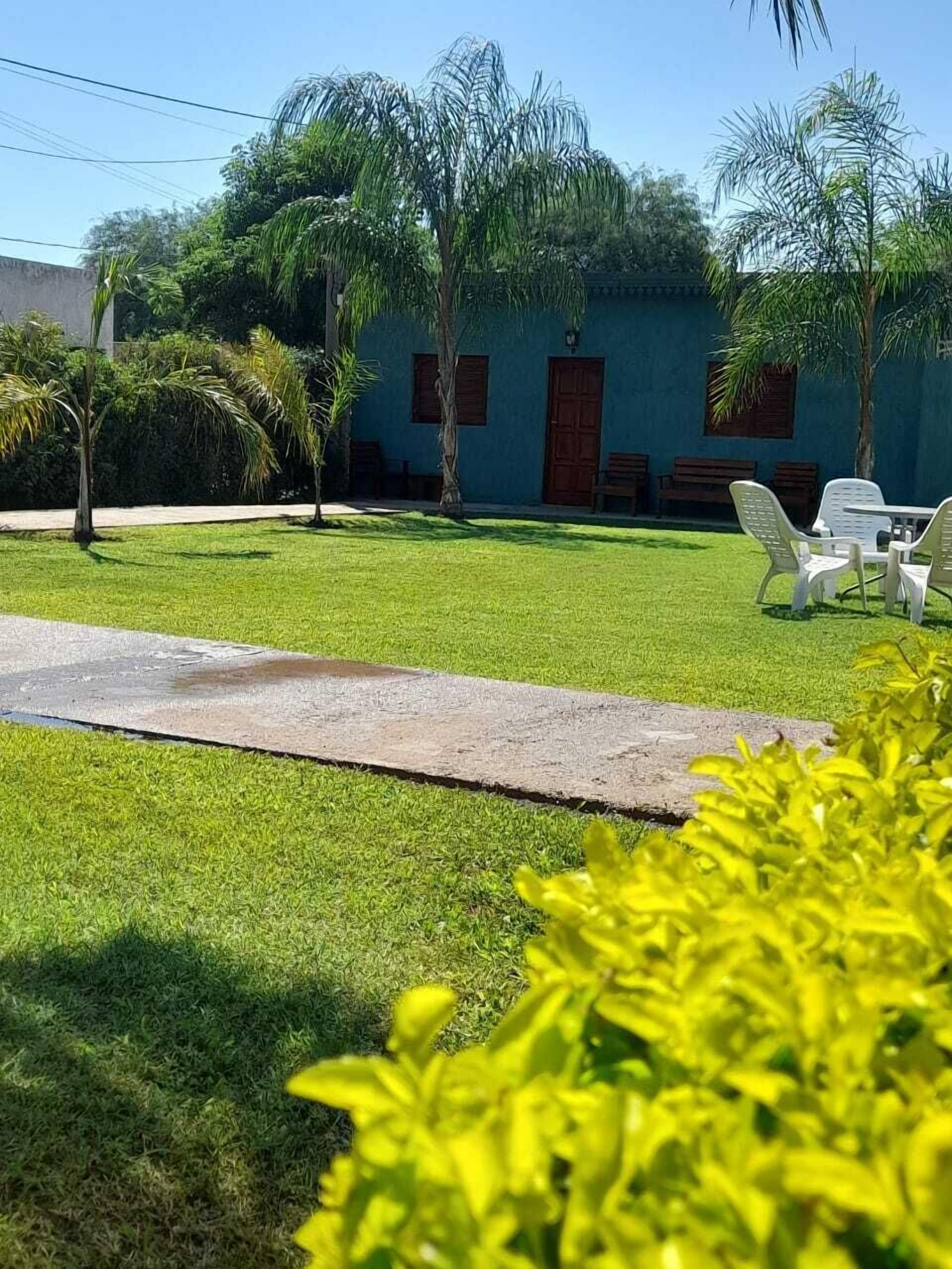 A well-maintained lawn with white plastic chairs and a concrete pathway leading to a building with a blue facade and wooden doors.