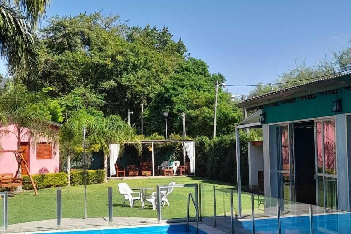 A swimming pool with a clear blue water, surrounded by a fenced area with lounge chairs and a small building with a green roof.