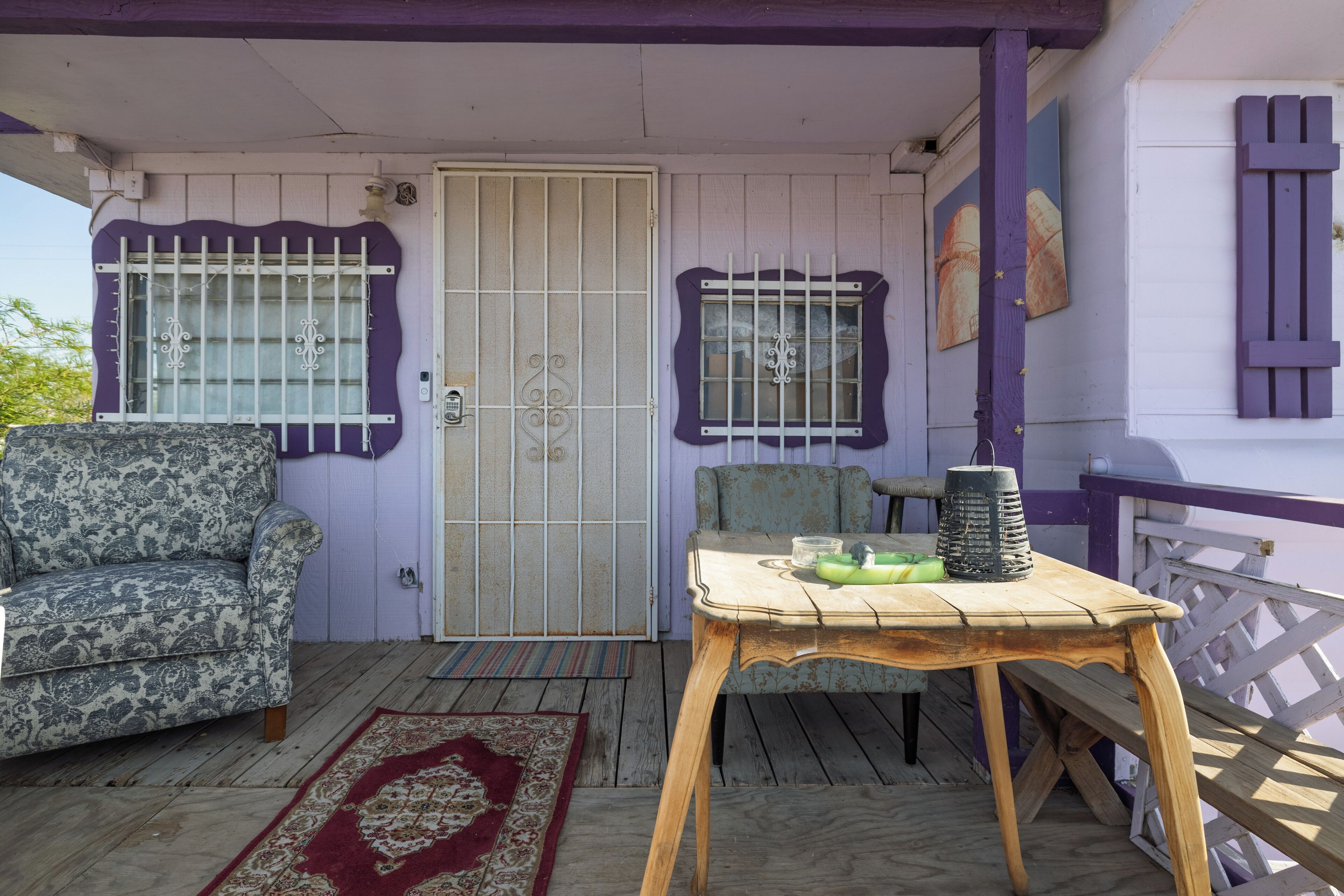 A porch with a wooden table, two chairs, a door with barred windows, and a railing.