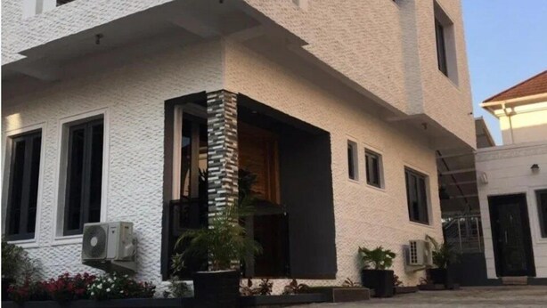 Un edificio de dos pisos con fachada blanca texturizada, un balcón con balaustrada de piedra y un amplio ventanal que refleja el cielo.