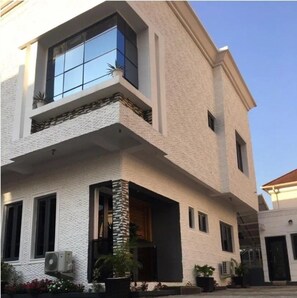 A two-story building with a white textured facade, a balcony with a stone balustrade, and a large window reflecting the sky. - Kodeland Apartment  (Lagos)