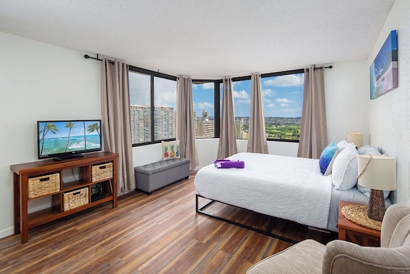 A hotel room with a large bed, a TV on a wooden console, a sofa, and a view of the city.