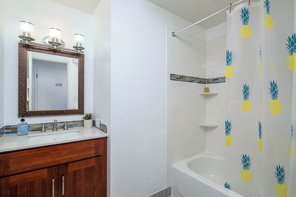 Une salle de bain avec un meuble-lavabo blanc, un miroir doté d’un cadre décoratif, une douche avec un rideau de douche à motifs d’ananas et une baignoire.