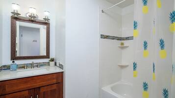 Une salle de bain avec un meuble-lavabo blanc, un miroir doté d’un cadre décoratif, une douche avec un rideau de douche à motifs d’ananas et une baignoire.