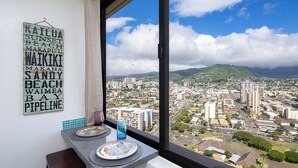 A dining area with a view of a cityscape through a large window, a wall-mounted sign with place names, and a table with plates and a glass. - Awesome view. Walking distance to beach, restaurants, shopping (Honolulu)