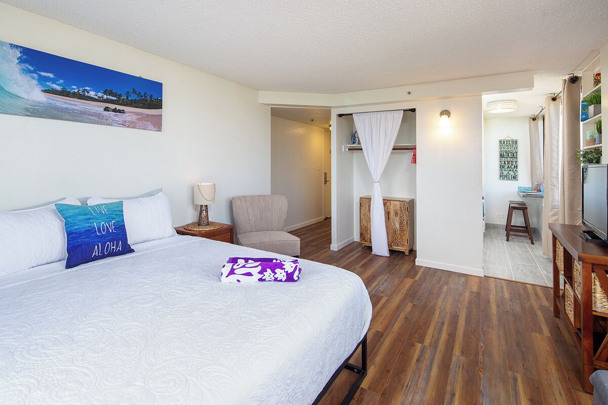 A hotel room with a large bed, a chair, a TV, and a view of the beach through a window.