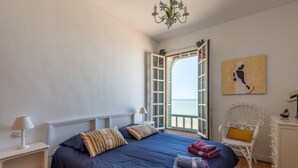 Un dormitorio con una cama grande, una mesita de noche blanca con una lámpara, una silla blanca con un cojín amarillo y un cuadro en la pared.