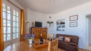 Un comedor con una mesa de madera, sillas, un jarrón con flores y una botella de champán. Se observa un televisor sobre un mueble, y un sofá marrón al fondo.