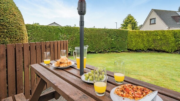 A wooden picnic table with food and drinks, a red umbrella, and a hedge in the background. - 3 bedroom accommodation in Lamlash (Lamlash)
