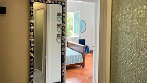 Een gang met een spiegel, een bed met een gestreept dekbedovertrek, en uitzicht op een kamer met een blauw gordijn.