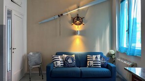 Een kamer met een blauwe bank, sierkussens met een nautisch thema, een wandlamp in de vorm van een stuurwiel en een raam met blauwe gordijnen.