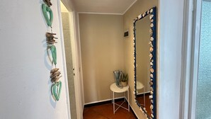 Een hal met decoratieve wanden, een spiegel met schelpen, een klein tafeltje met planten en een betegelde vloer.