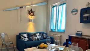 Een eetgedeelte met een tafel gedekt voor twee personen, een blauwe bank met kussens met nautische motieven, een aan de muur bevestigde scheepslamp, een raam met blauwe gordijnen en een klok aan de muur.