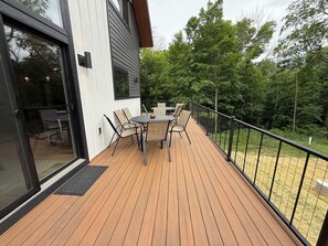 Una terraza de madera con una mesa y sillas, rodeada por una barandilla de metal, y un ventanal de vidrio que da a un espacio interior.