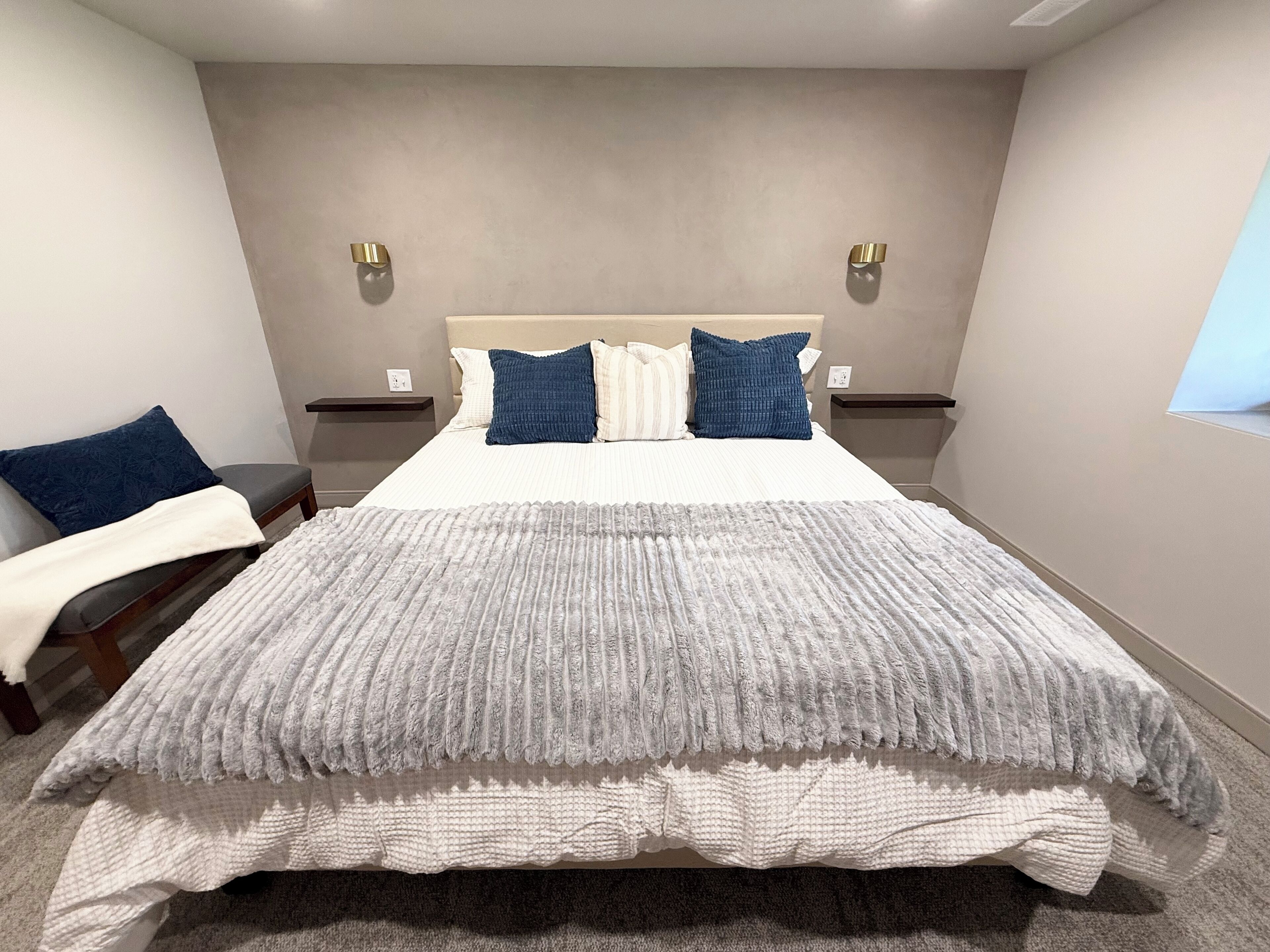A modern bedroom with a large bed, two bedside tables, a nightstand, a window, and a wall-mounted light fixture.