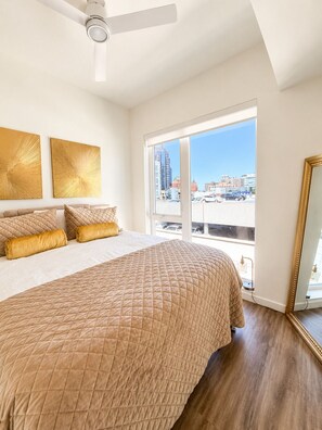 Un dormitorio con una cama grande, un ventilador de techo, un ventanal con vistas a la ciudad y dos obras de arte colgadas en la pared.