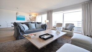 A modern living room with a grey sofa, a coffee table, and a beige armchair. There is a large window with curtains, a painting on the wall, and a lamp on the coffee table. - Apartment in Duinbergen by the Seafront (Knokke-Heist)