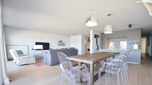 A modern living room with a wooden dining table and clear plastic chairs, a grey sofa, and a flat-screen TV. - Apartment in Duinbergen by the Seafront (Knokke-Heist)