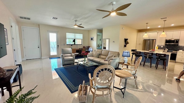 A spacious living area with a sofa, armchairs, a coffee table, and a dining area with a bar. There are ceiling fans, a kitchen with modern appliances, and a large window providing natural light.