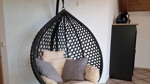 Un fauteuil suspendu doté d’une structure en treillis et de coussins, un sol carrelé et un meuble noir adossé à un mur blanc.