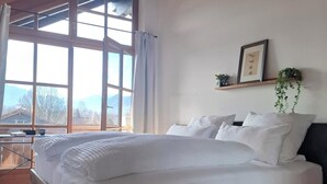 Une chambre avec un lit en bois, du linge de lit blanc, une grande fenêtre avec des rideaux, une étagère avec des plantes et un tableau encadré au mur.