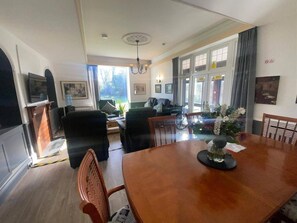 A dining area with a wooden table, chairs, and a vase with flowers. There are windows with curtains, a television, and framed pictures on the walls. - 6 Bed-sleeps 8 Gym-hottub-garden-parking (Bristol)