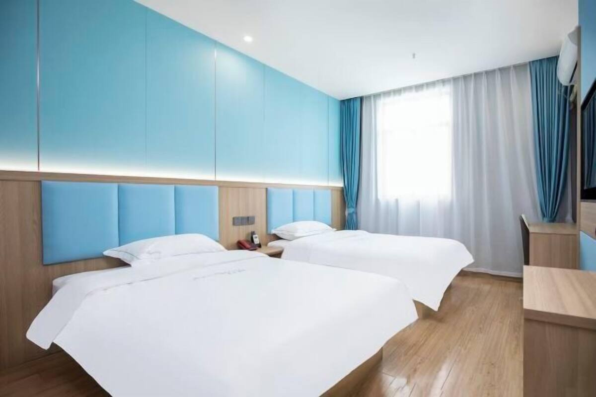 A hotel room with two beds, a wooden headboard, a bedside table, and a large window with curtains.