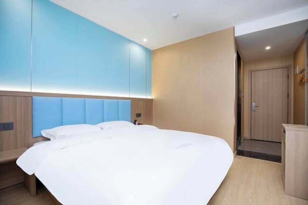 A modern hotel room with a large bed, a built-in headboard, and a wooden wardrobe.