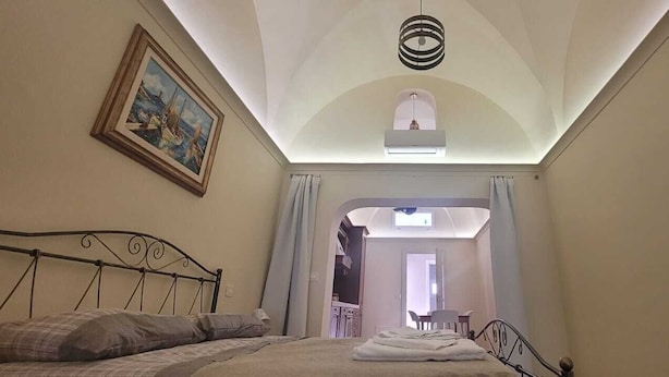 Ein Schlafzimmer mit Bett, einem Gemälde, einer Deckenleuchte und Blick in einen anderen Raum.