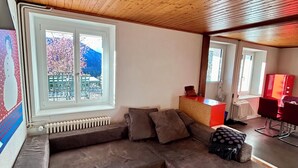 Una sala de estar acogedora con techo de madera, un ventanal con vistas a los cerezos en flor, un sofá seccional y una pequeña área de cocina.