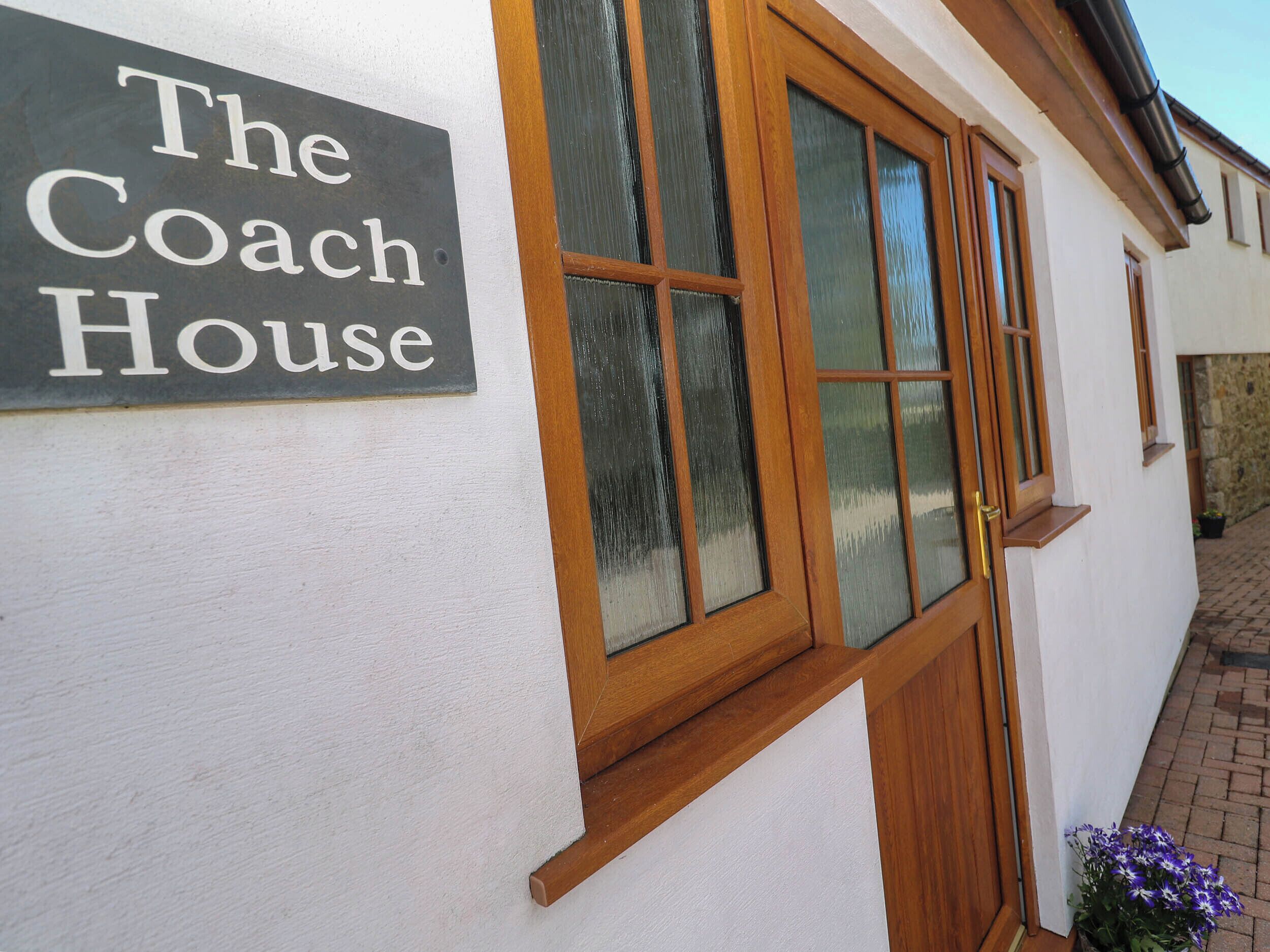 Un letrero que dice 'The Coach House' sobre una pared blanca, ventanas con marcos de madera y una pequeña maceta con flores moradas.