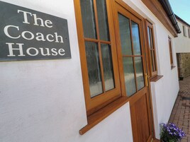 Un letrero que dice 'The Coach House' sobre una pared blanca, ventanas con marcos de madera y una pequeña maceta con flores moradas.