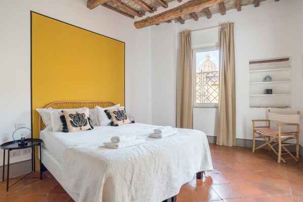 A bedroom with a large bed, a bedside table, a chair, a window with a view of a building, and a shelf with items.