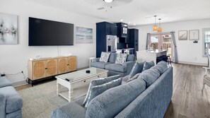 Una sala de estar moderna con un sofá gris, un mueble de madera, un televisor de pantalla plana y una zona de cocina con gabinetes oscuros.