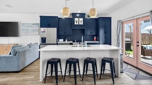 Une cuisine moderne avec des meubles de couleur bleu foncé, un îlot central blanc doté de tabourets de bar et un grand téléviseur fixé au mur.