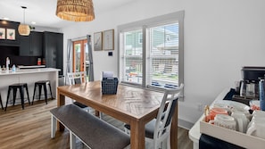 A modern kitchen with a wooden dining table, bar stools, and a bench. There are two windows with blinds, a hanging light fixture, and a kitchen counter with appliances.