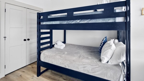 Una cama litera con colchón gris y almohadas blancas, una puerta blanca y un suelo de madera.