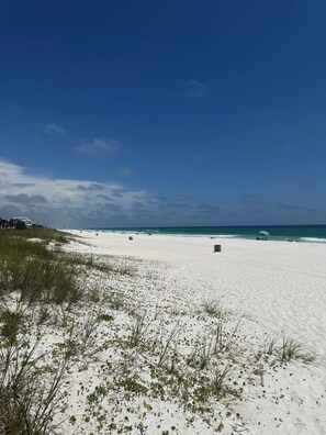 Una playa de arena blanca, vegetación escasa y cielo azul despejado.