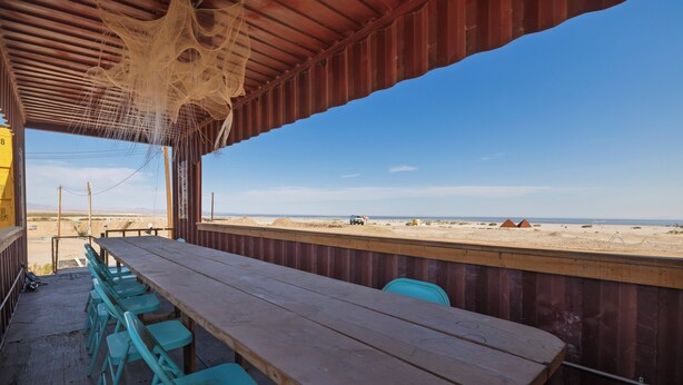 Un espace de restauration en plein air avec une longue table en bois et des chaises turquoise, donnant sur une plage.