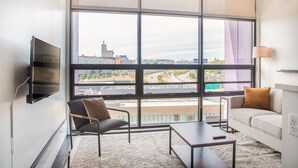 Ett modernt hotellrum med ett stort fönster som erbjuder utsikt över staden, en platt-TV som är monterad på väggen, en soffa, en fåtölj och ett litet bord.