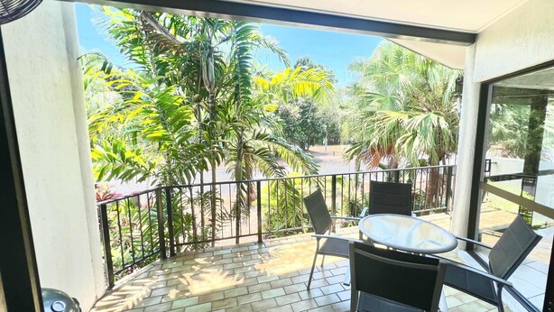 A balcony with a table and chairs, surrounded by a metal railing, overlooking a garden with various trees and plants.