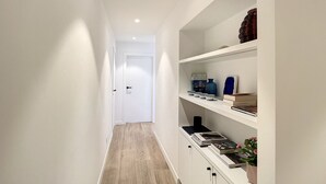 A narrow hallway with wooden flooring, white walls, and a white door at the end. Shelves on the right hold books, a vase, and a plant. - Apartment in Seaside Bliss (Knokke-Heist)