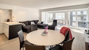 A modern living room with a sofa, a dining table, and a kitchen area. - Apartment in Seaside Bliss (Knokke-Heist)