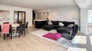 A modern living room with a sectional sofa, a dining area with a table and chairs, and a kitchen in the background. - Apartment in Seaside Bliss (Knokke-Heist)