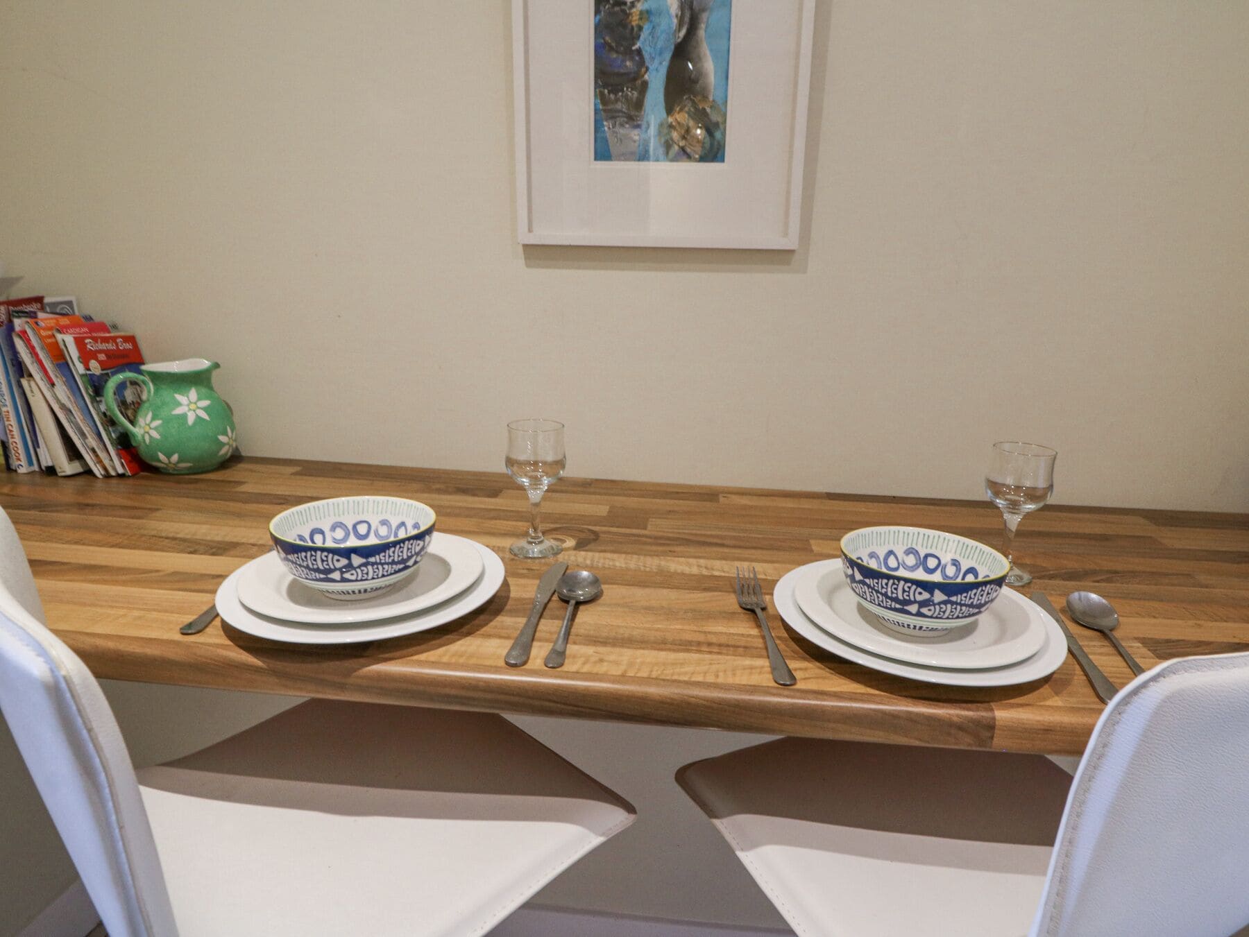Une table à manger dressée avec des bols, des assiettes, des couverts et des verres, ainsi que des magazines et un pichet vert posé à côté.
