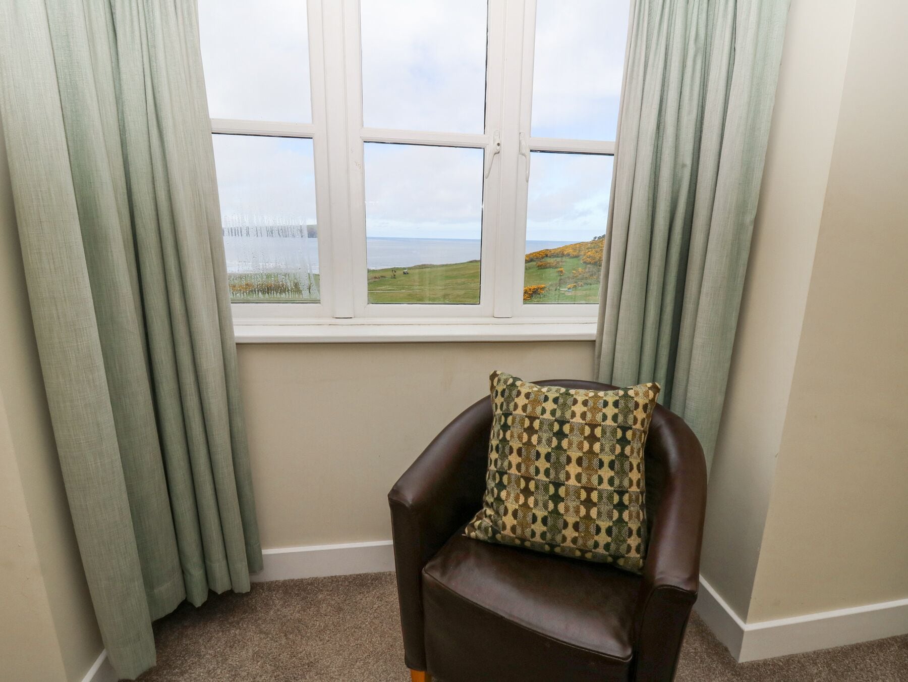 A corner room with a view of the sea through a window, a brown leather chair, and light green curtains.