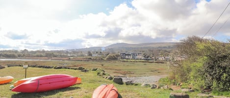 A grassy area with two kayaks, a rocky terrain, and a distant view of houses and hills under a partly cloudy sky.